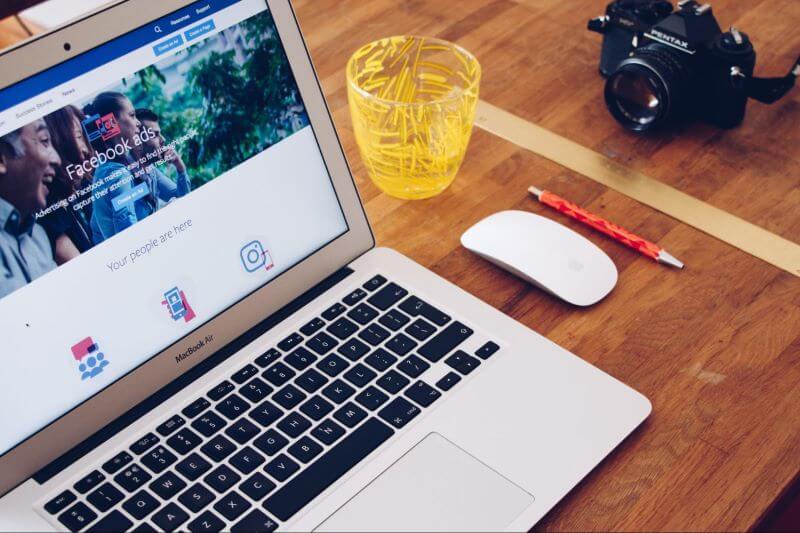 A laptop sits open on a desk adjacent to a mouse, pen, glass and camera. The screen shows the Facebook ads homepage as part of the article, ‘How does Facebook analytics work?